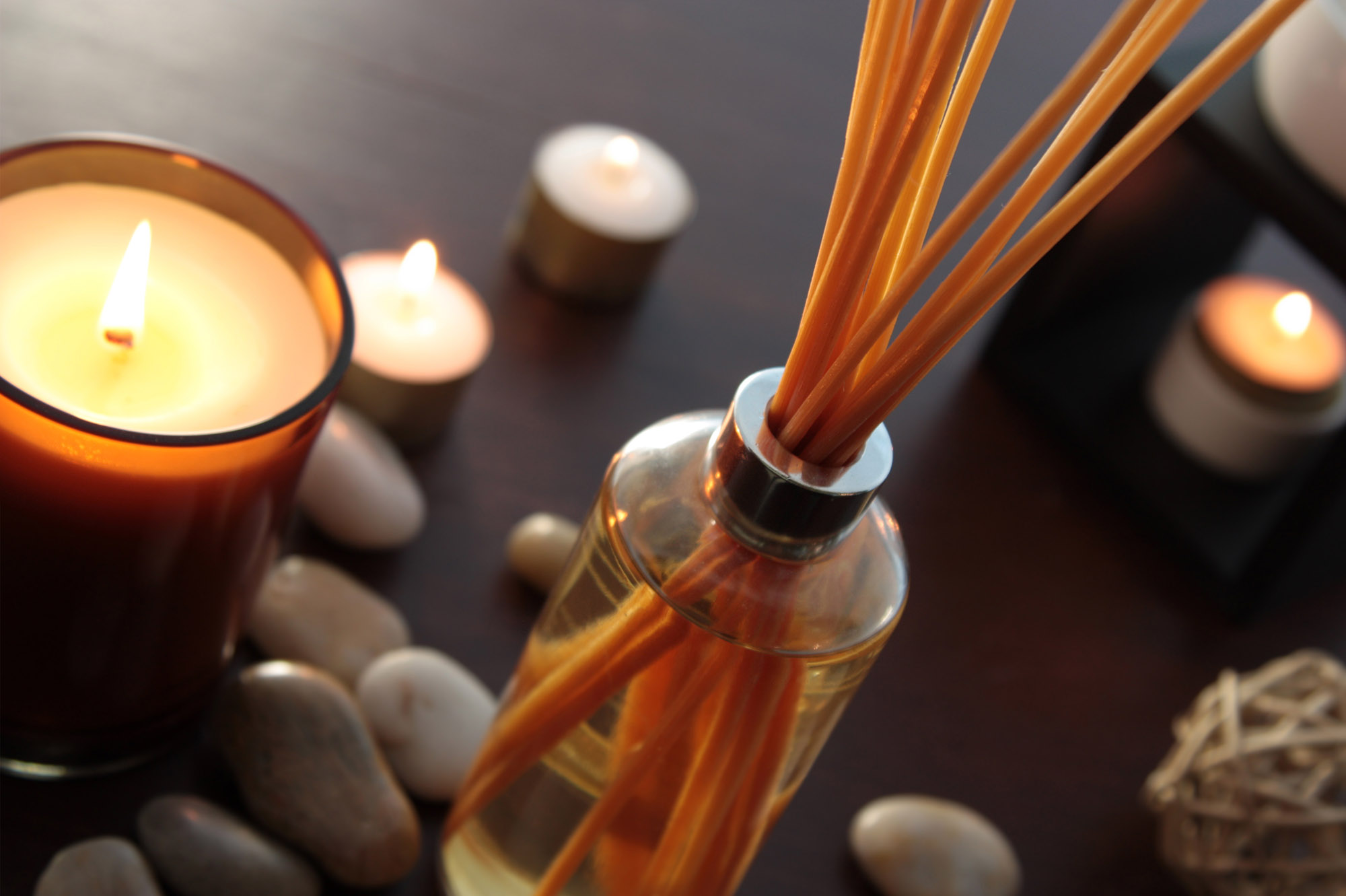 Close up of a bottle of fragrance diffuser with reeds. Tea light candles and an oil burner in the background. Shallow depth of field.